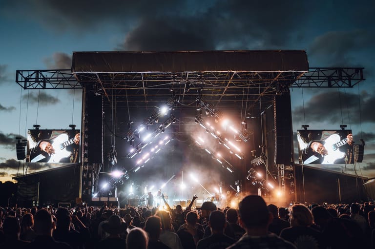 Large outdoor concert stage with dramatic lighting and pyrotechnics, surrounded by thousands of audience members with raised hands.