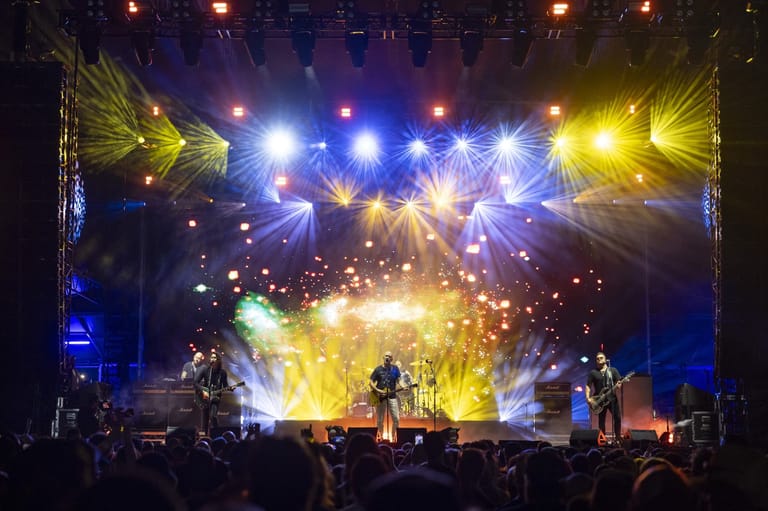 Band performing on stage with dramatic blue and yellow lighting, large wing-shaped set pieces, and crowd silhouettes in foreground.