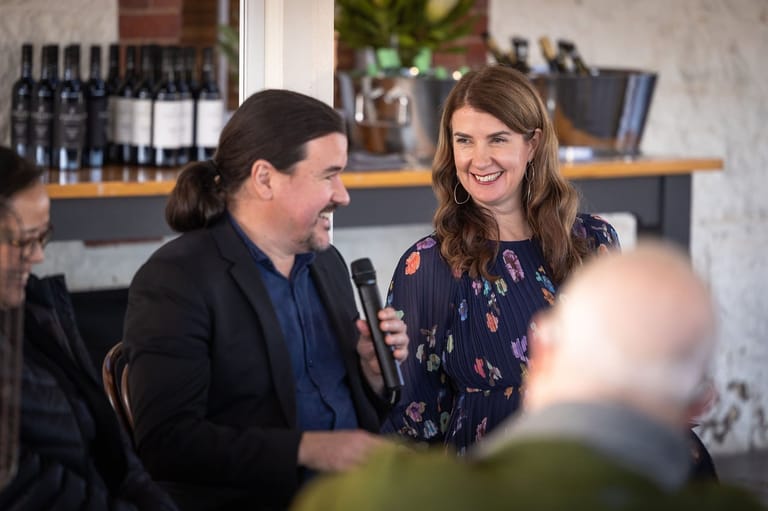 Two people speak into microphones at an indoor event with wine bottles displayed on shelves behind them.