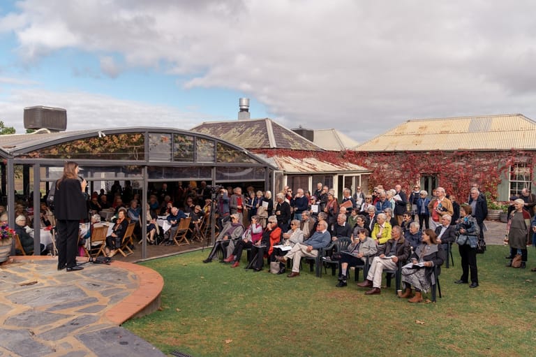 Outdoor event with crowds seated and standing on manicured lawns beside heritage brick buildings under partly cloudy sky.