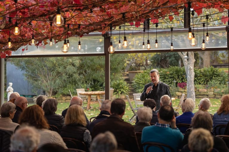 Speaker addresses seated audience beneath red-leafed pergola with hanging lights and garden views.