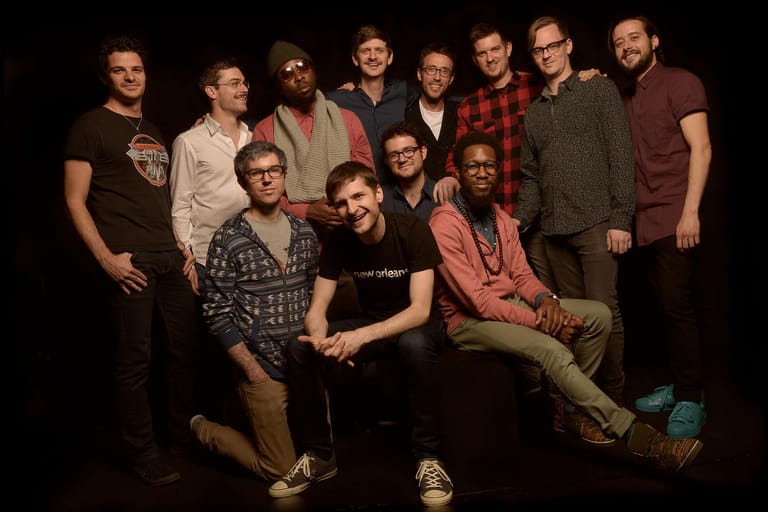 A group of nine musicians pose together against a dark background in casual clothing.