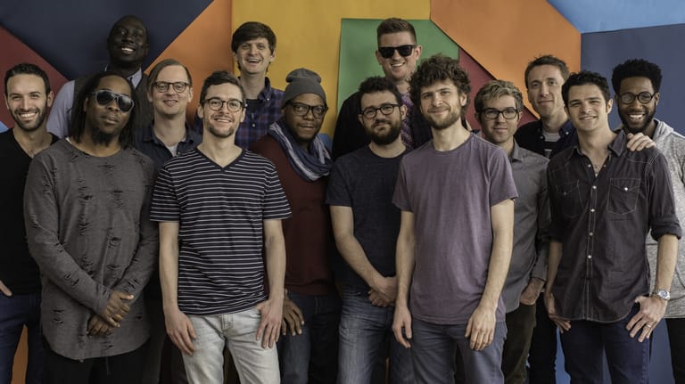 Large group of musicians posed together against a colourful geometric backdrop indoors.