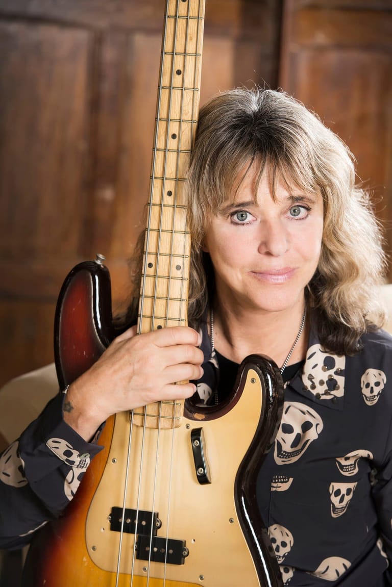 Musician with blonde hair holding a bass guitar, wearing a black shirt with skull patterns.