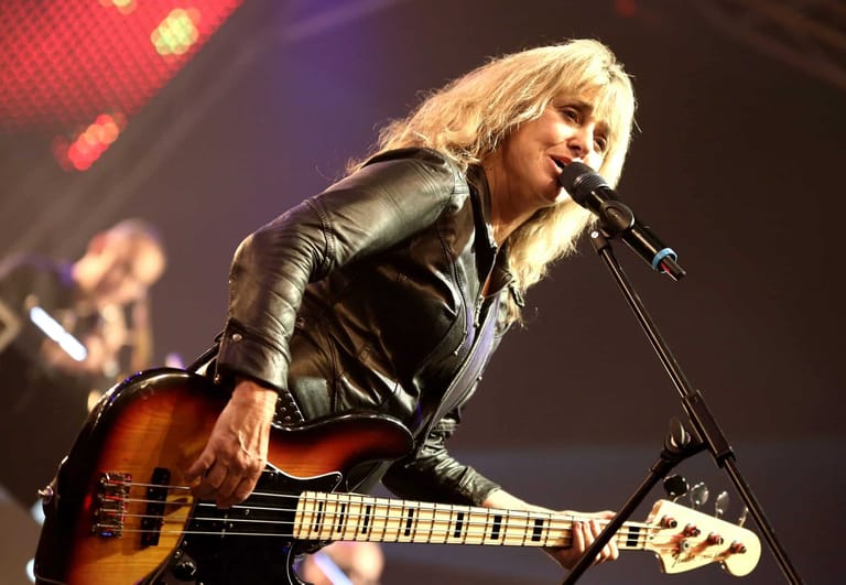 Musician with platinum blonde hair performs at microphone whilst playing acoustic guitar under warm stage lighting.