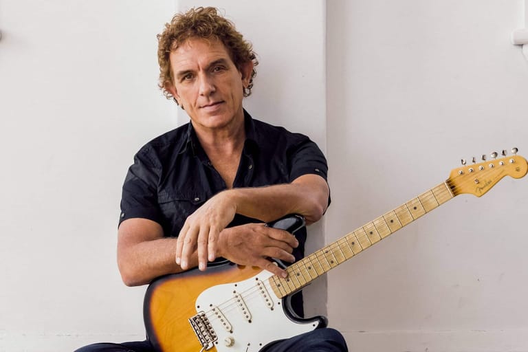 Musician with curly hair holding a yellow electric guitar against a white wall.