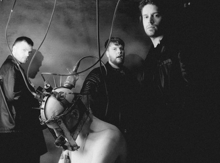 Three musicians pose with vintage microphones and studio equipment in a moody black and white photograph.