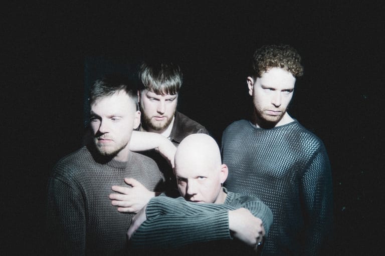 Four young men in grey knit sweaters posed together against a dark background.