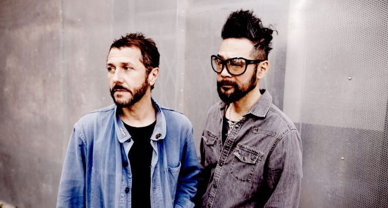 Two men stand against a grey wall, one wearing a blue shirt and the other in a grey jacket and glasses.
