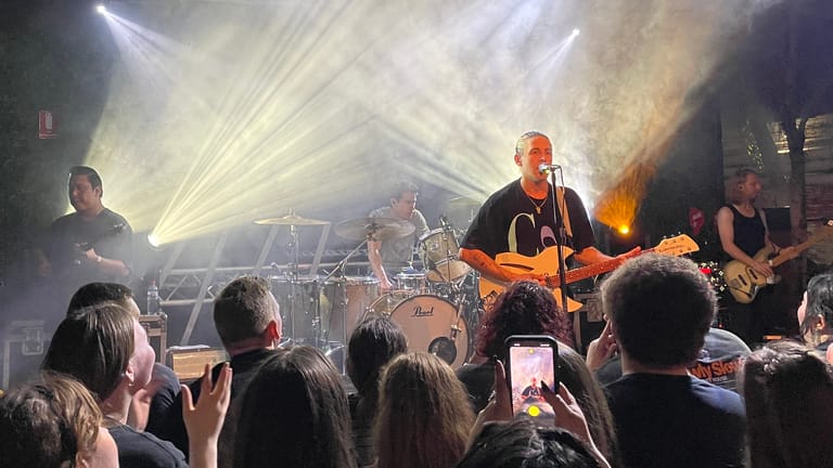Musician in orange shirt performing on stage with band, dramatic lighting and crowd in foreground.