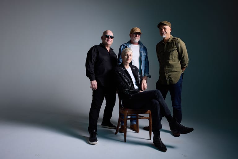 Four musicians posed together against a grey studio backdrop, one seated on a wooden chair and three standing.