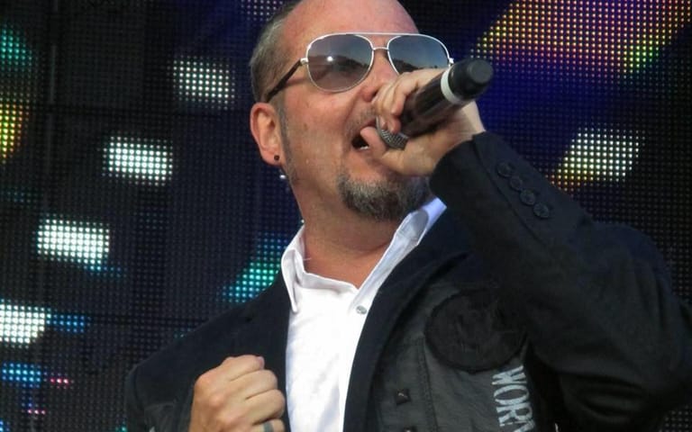 Performer in sunglasses and dark jacket singing into microphone against illuminated LED screen backdrop.