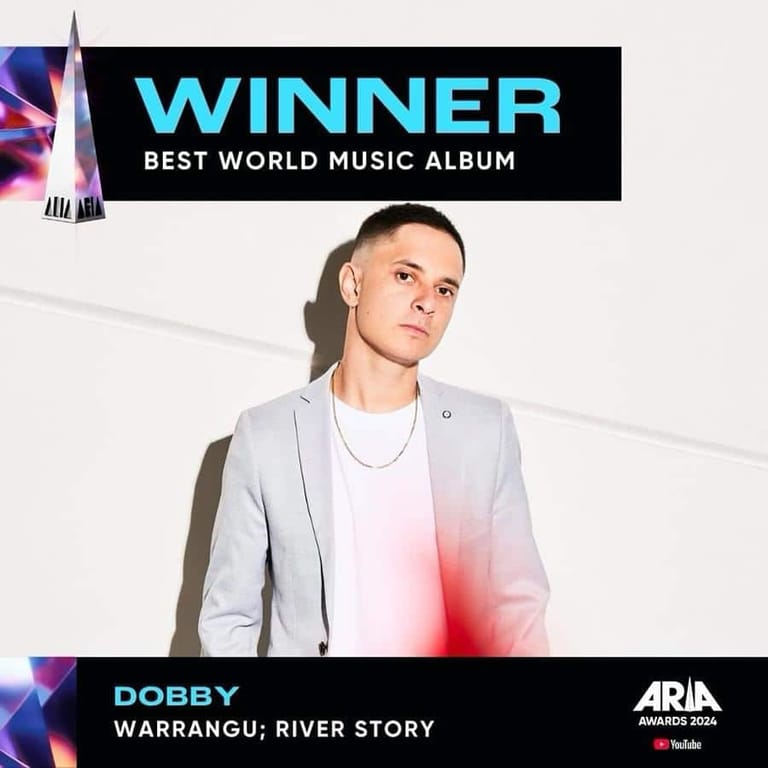 Dobby poses in a light grey blazer against a white background, alongside a banner announcing his album "Warrangu; River Story" as winner of Best World Music Album at the ARIAs.