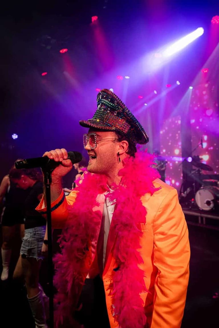 Performer in flamboyant pink and yellow outfit with studded cap sings at microphone under vibrant purple and magenta stage lighting.