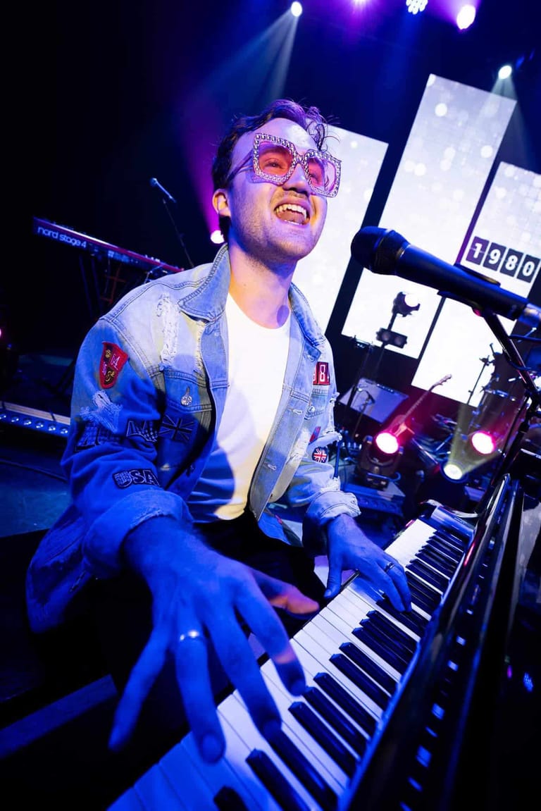 Performer in pink-tinted glasses and denim jacket plays keyboard under blue stage lighting during a theatre production.