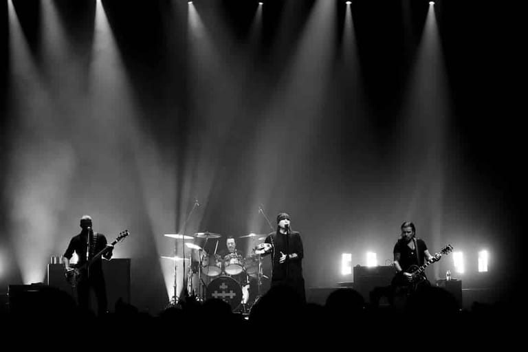 Four-piece band performing on stage under dramatic white spotlights in black and white.