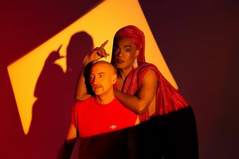 Two performers in red lighting against a dark backdrop with dramatic yellow stage lighting and shadows.