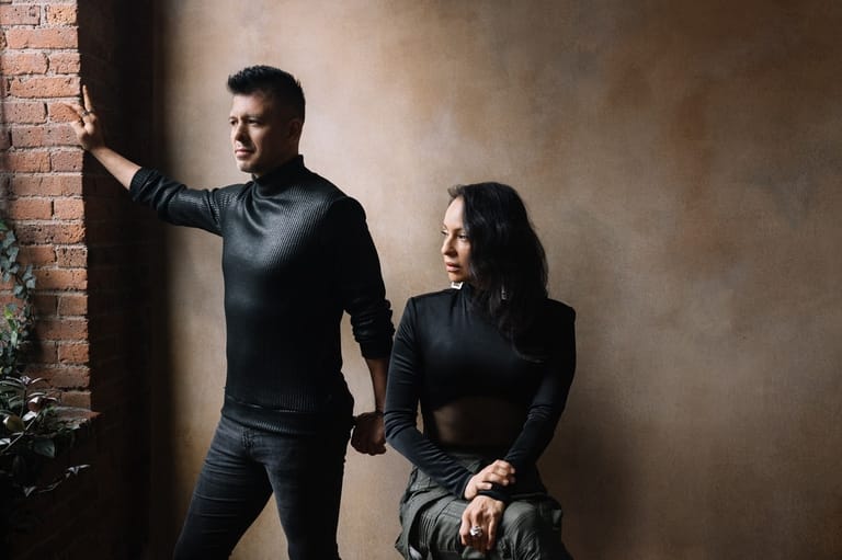 Two performers in black clothing pose against a brick wall in dramatic studio lighting.