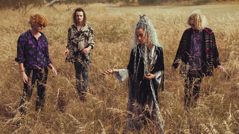 Four musicians stand in a sparse, dry grassland wearing dark clothing.