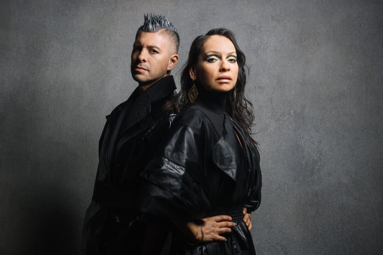 Two musicians in black leather jackets pose back-to-back against a grey studio backdrop.