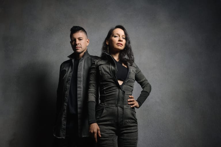 Two performers in black leather jackets stand confidently against a grey studio backdrop.