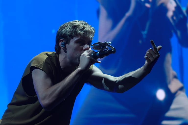 Performer singing into microphone on stage bathed in blue light during The First Time Tour.