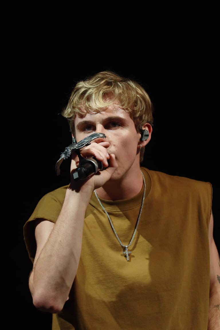 Performer singing into microphone against black background, wearing mustard-yellow sleeveless shirt and cross pendant necklace.