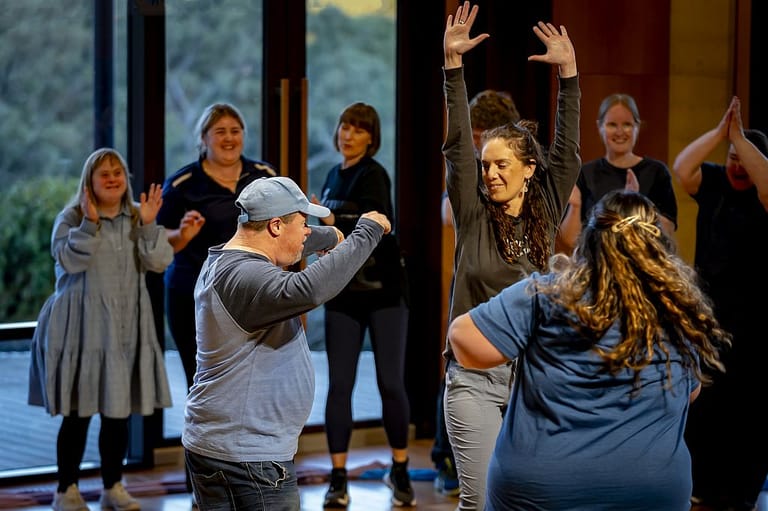 Group of people participating in an interactive workshop or rehearsal, with some raising their arms and engaging with facilitators in a theatre space.