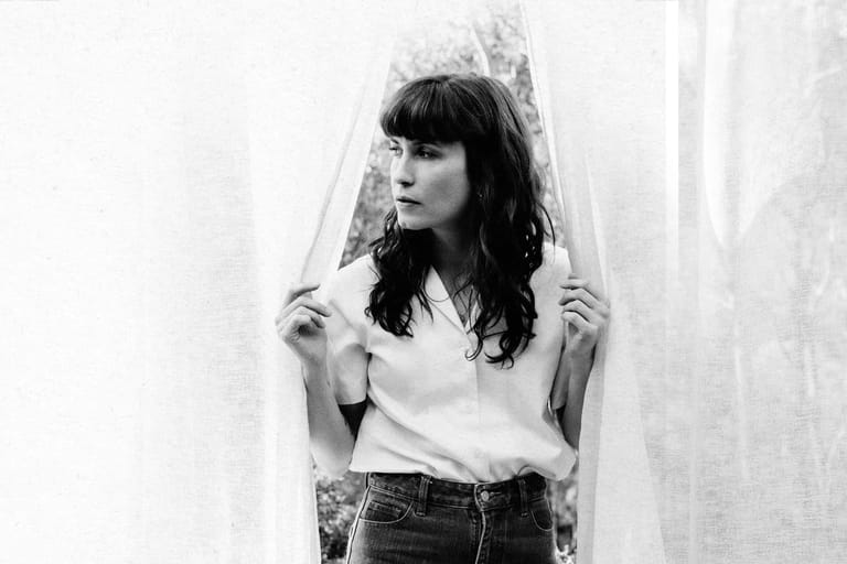 Woman with dark wavy hair holding sheer curtain fabric in black and white portrait.