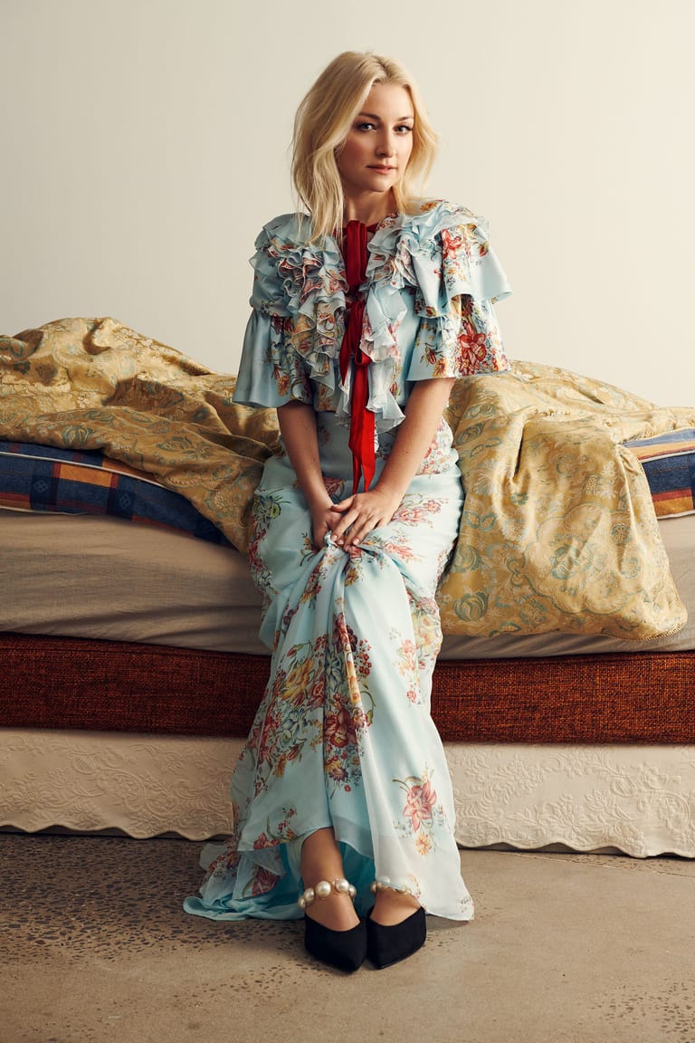 Woman in a floral dress and layered cardigan poses against an ornate headboard in a styled interior setting.