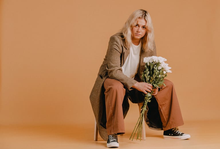 Woman with long blonde hair sitting cross-legged holding white flowers against warm beige background.
