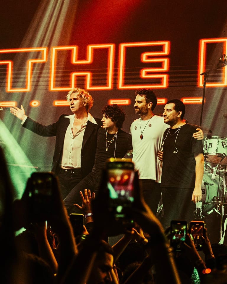 Four band members on stage beneath glowing neon lettering, with audience members filming in the foreground.