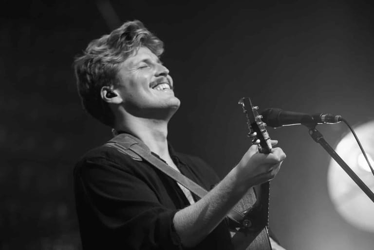 Musician performing on stage with acoustic guitar and headset microphone under dramatic spotlight.
