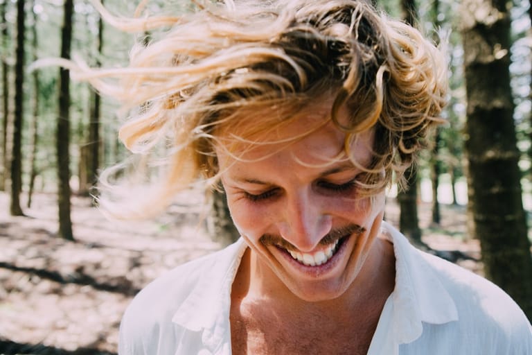 Person with blonde hair smiling outdoors in a sunlit forest setting.
