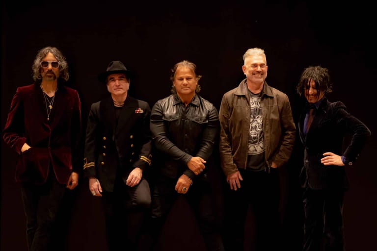 Five musicians in dark clothing stand in a line on stage against a black background.