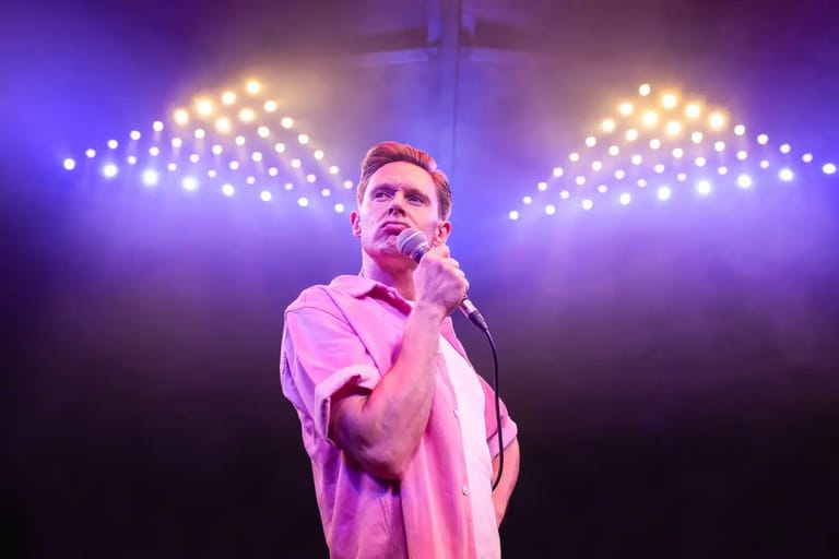 Male performer in pink shirt singing into microphone under purple stage lighting with geometric light display.