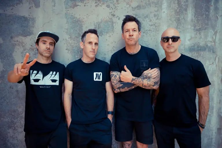 Four men in black t-shirts stand against a weathered grey wall in a band promotional photo.