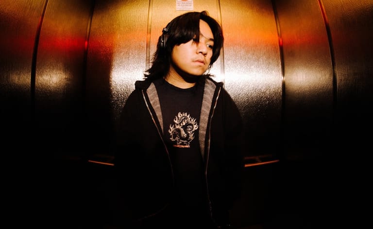 Person in dark hoodie stands against warm-lit metallic backdrop with orange and amber lighting.