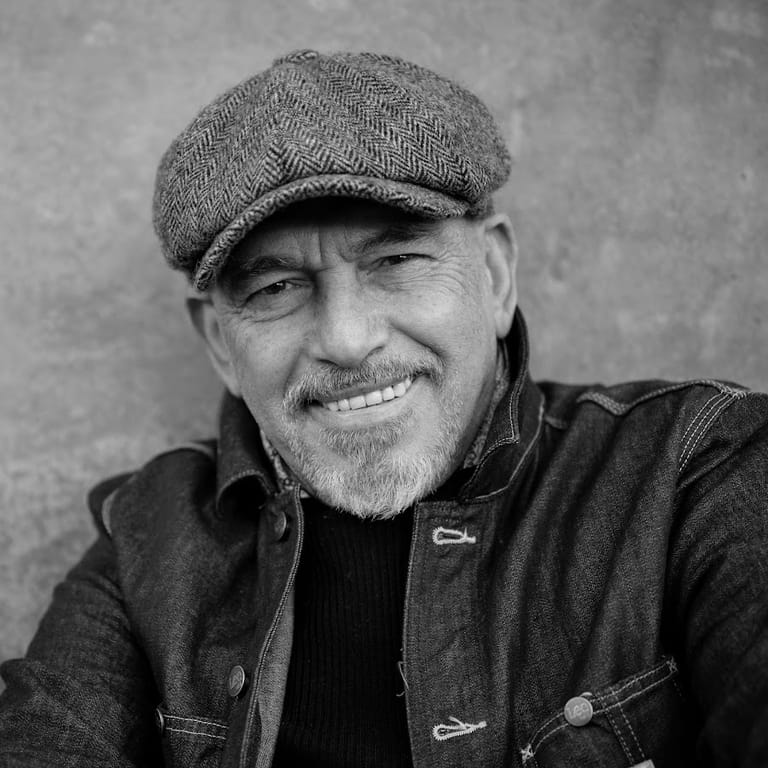 Man wearing a grey flat cap and dark jacket smiles at the camera in a black and white portrait.