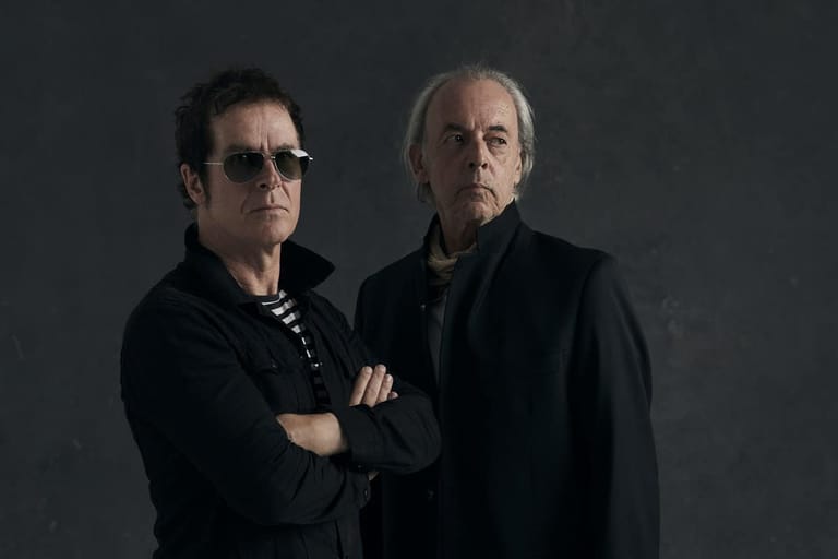 Two musicians in dark clothing posed against a grey background, one wearing sunglasses with arms crossed.