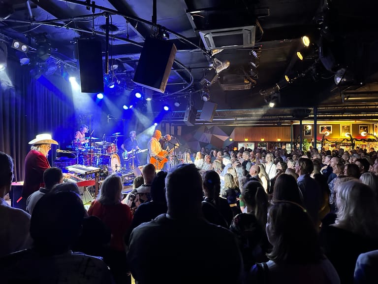 Band performing on stage with blue and warm lighting while audience members watch from the floor in an indoor venue.