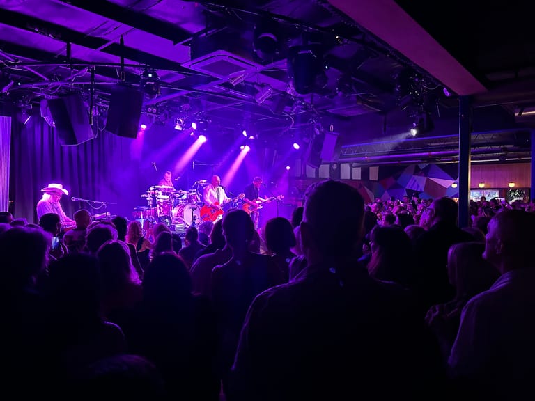 Band performs on stage bathed in purple lighting as audience members watch from the floor.