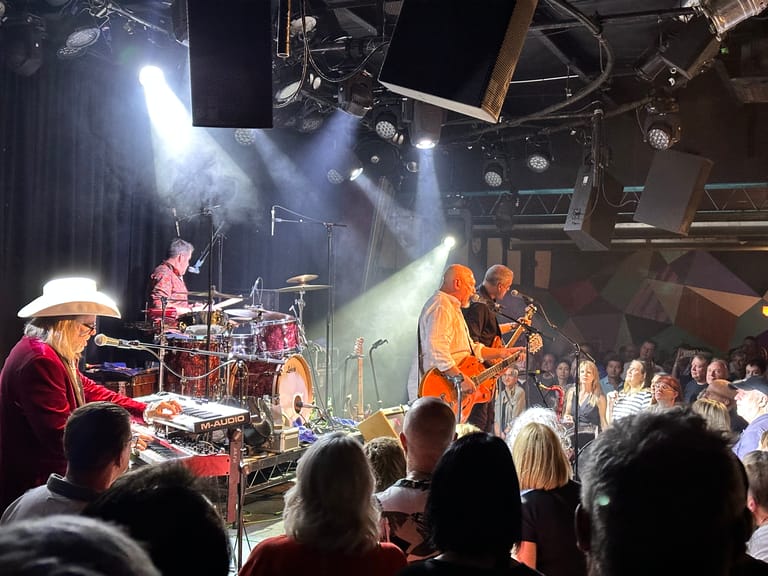 Musicians perform on stage under dramatic lighting, with drummer and band members visible amid audience members in foreground.