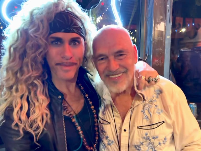 Two performers posing together at a venue, one wearing a headband and dark clothing, the other in a cream patterned shirt.