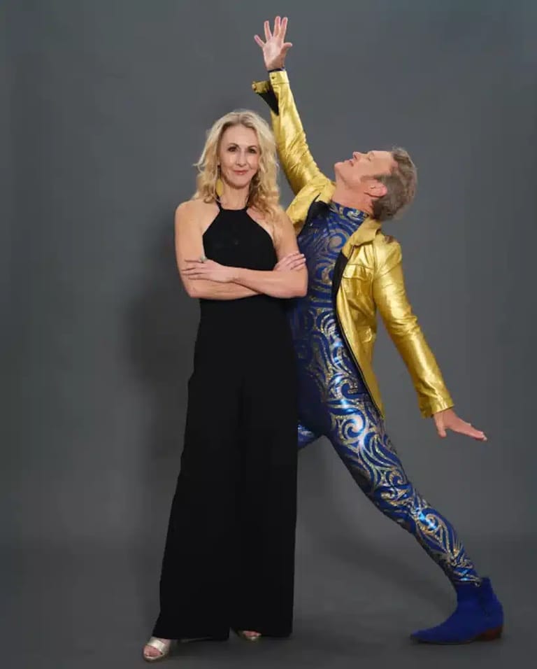 Two performers pose against a grey backdrop, one in a black dress with arms crossed, the other in a gold and blue sequined costume striking a dramatic lean-back pose.
