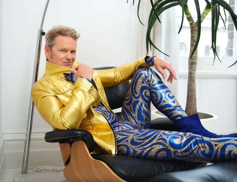 Man in gold jacket reclines in chair wearing patterned blue and gold trousers against white wall with potted plant.