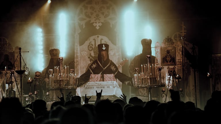 Theatrical stage production with dramatic lighting, gothic architecture, and silhouetted audience members in foreground.