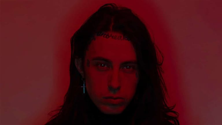 Person with long dark hair against a deep red background lit by moody stage lighting.