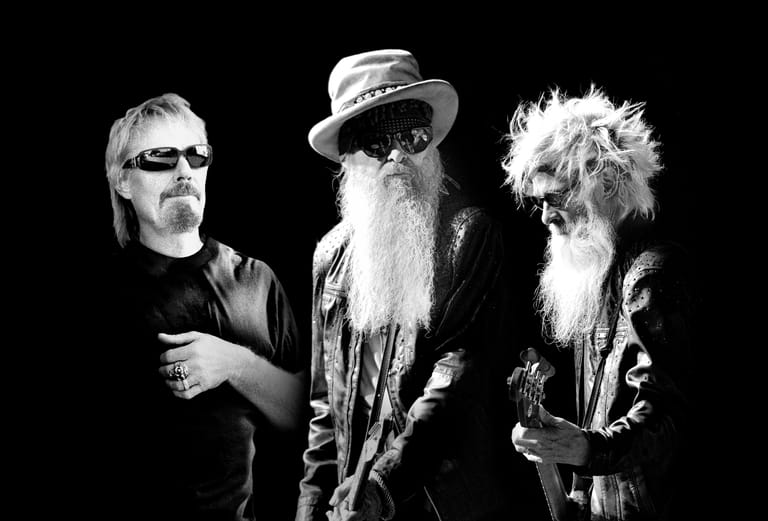 Three musicians in black and white, featuring two with long beards, one wearing a wide-brimmed hat, posed against a dark background.