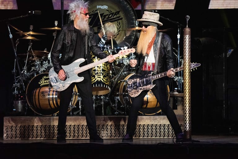 Two guitarists perform under stage lights, one in a black jacket with white hair, the other wearing a hat and sunglasses.
